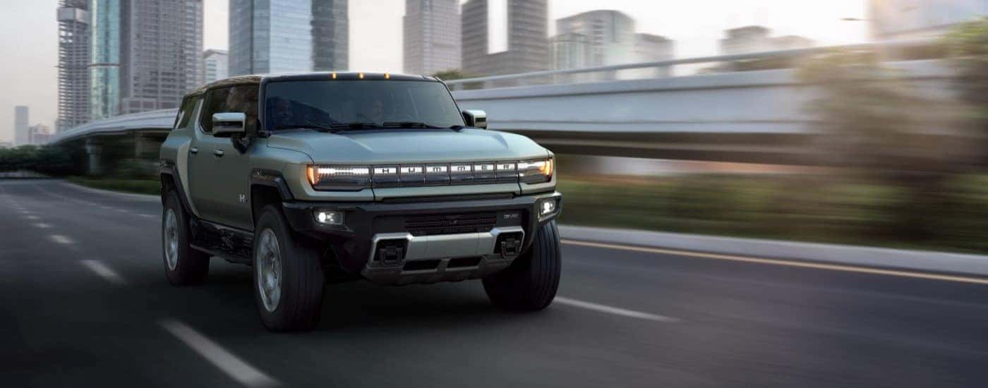 A green 2024 GMC Hummer EV is shown driving past city buildings.