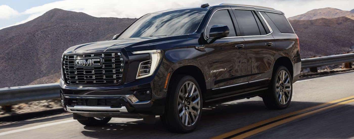 A black 2026 GMC Yukon Denali driving down a road with mountains in the background.