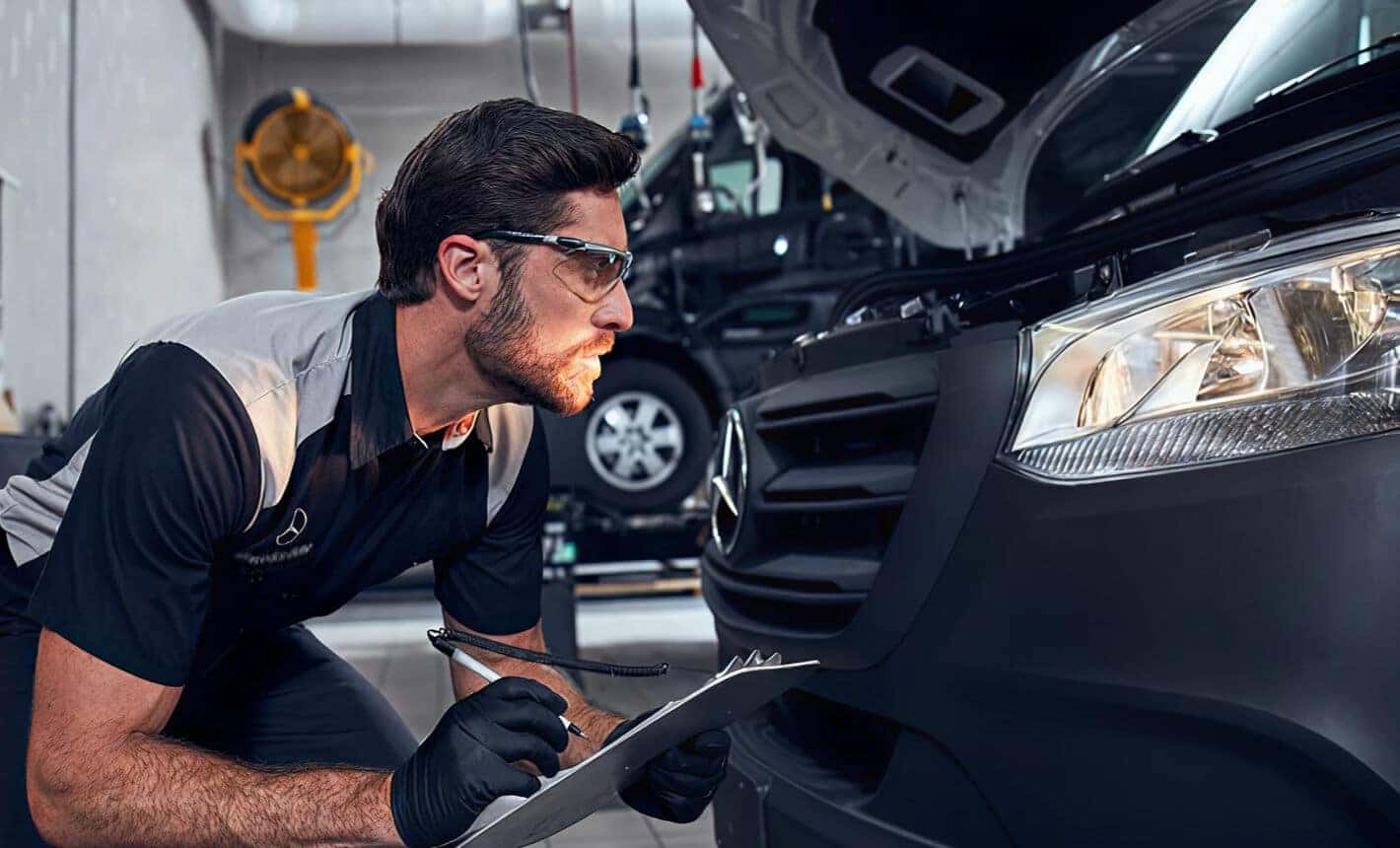 A Mercedes-Benz Technician inspects the headlamps on a Mercedes car while taking notes on a clipboard.