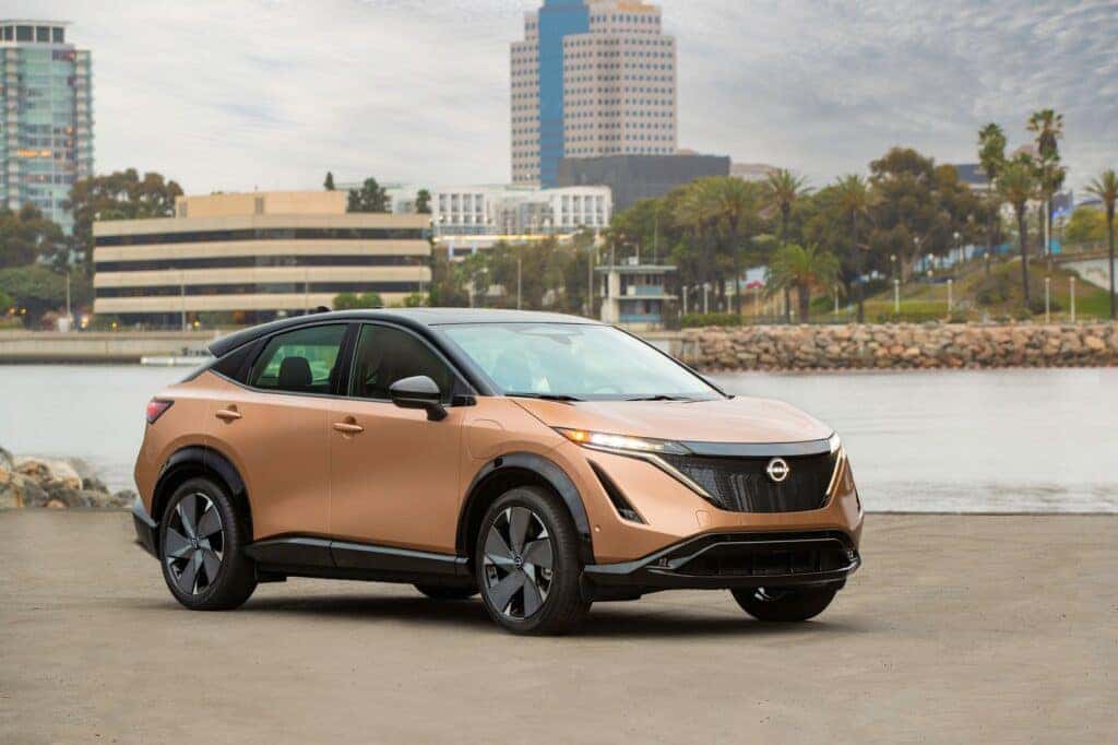 A Gold And Gorgeous 2024 Nissan Ariya Parked By A Lake, A Cityscape In The Background