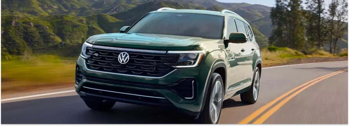 Front three-quarter view of a green 2026 Volkswagen Atlas driving on a paved road with mountains in the background.